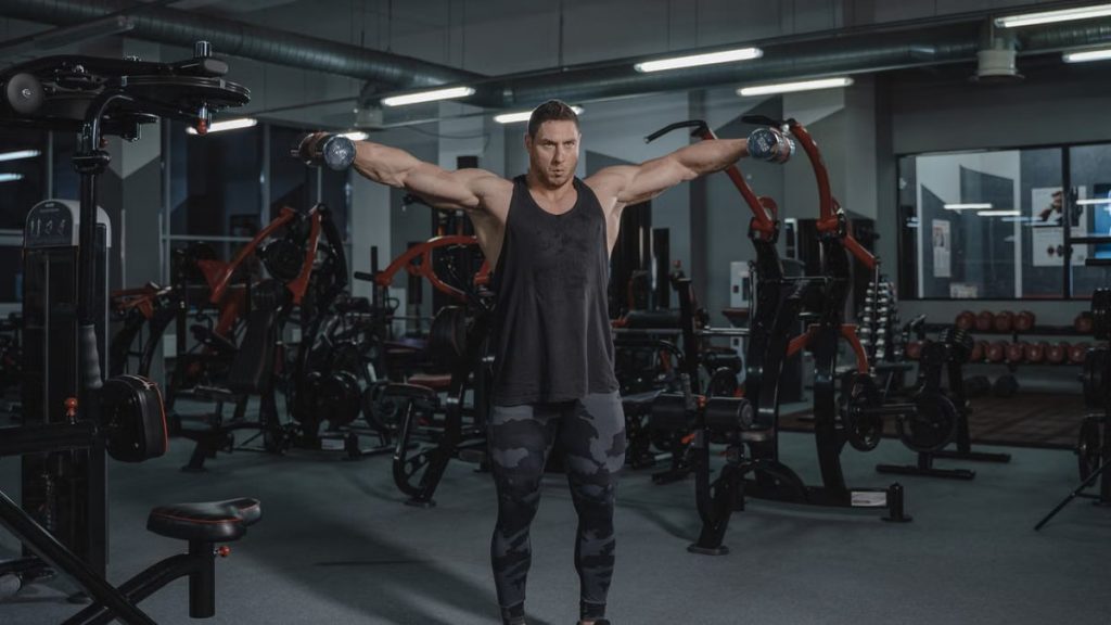 A muscular man in a black tank top performing dumbbell raises in a modern gym.